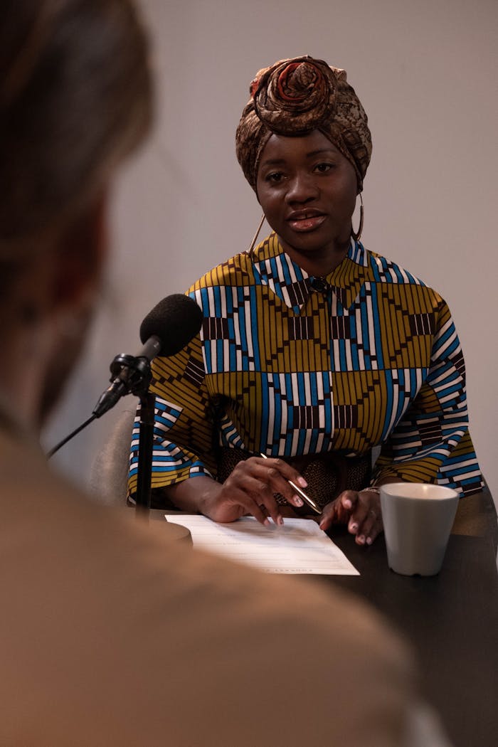 Black woman in vibrant attire hosting a podcast interview, talking into a microphone.